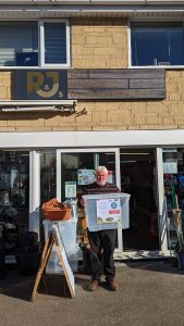 A man standing outside a shop is holding a clear, plastic box which people can donate items like canned food in, for people struggling with the cost of living,