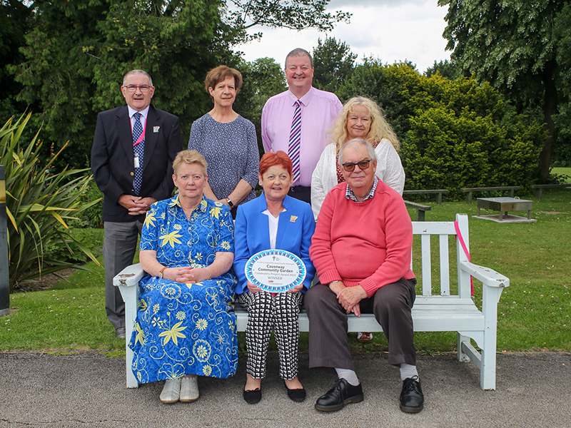 Recipients of the 2023 Community Project Awards, standing behind and sitting on a bench in the grassy grounds of The Hayes Conference Centre, with Andy Jackson, Head of Communications who wears a pink shirt.
